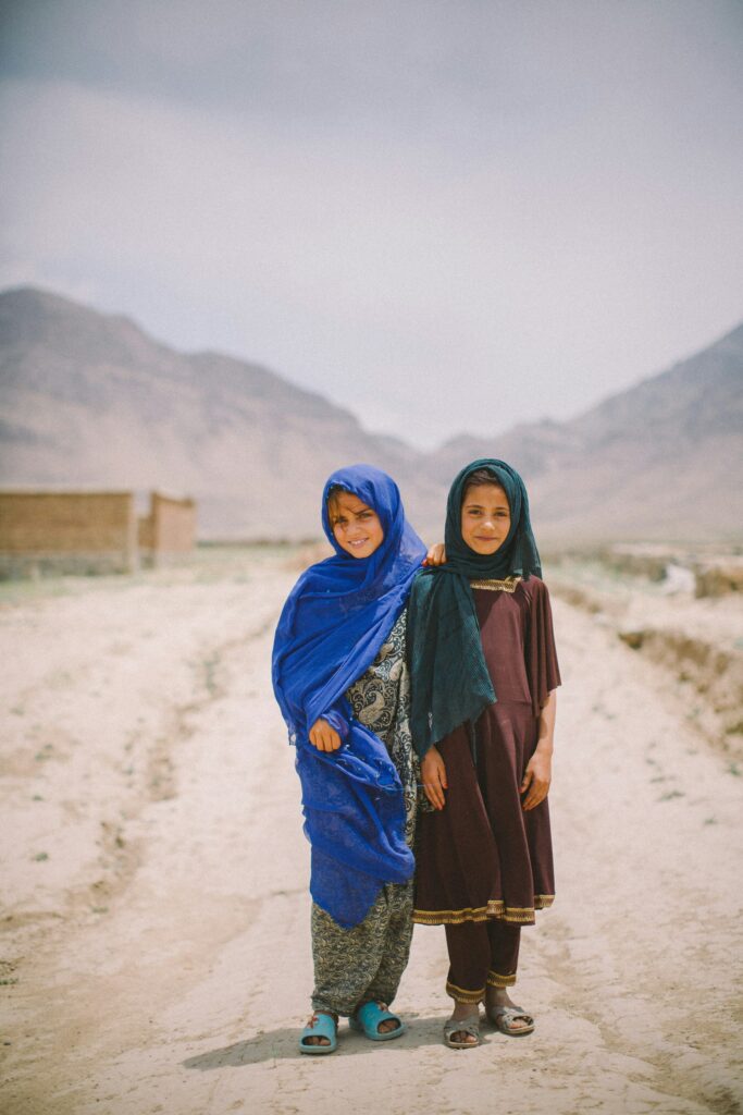 Two young Afghan girls stand on a sandy road